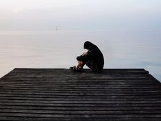 Photo of an individual in a hooded sweatshirt sitting and holding their knees with head bent down at the end of a long wooden pier or dock. A large expanse of water is in the background. Person is in distress, isolated, alone, struggles with ED and low sexual desire and could benefit from sex therapy, in the Minneapolis, Minnesota area