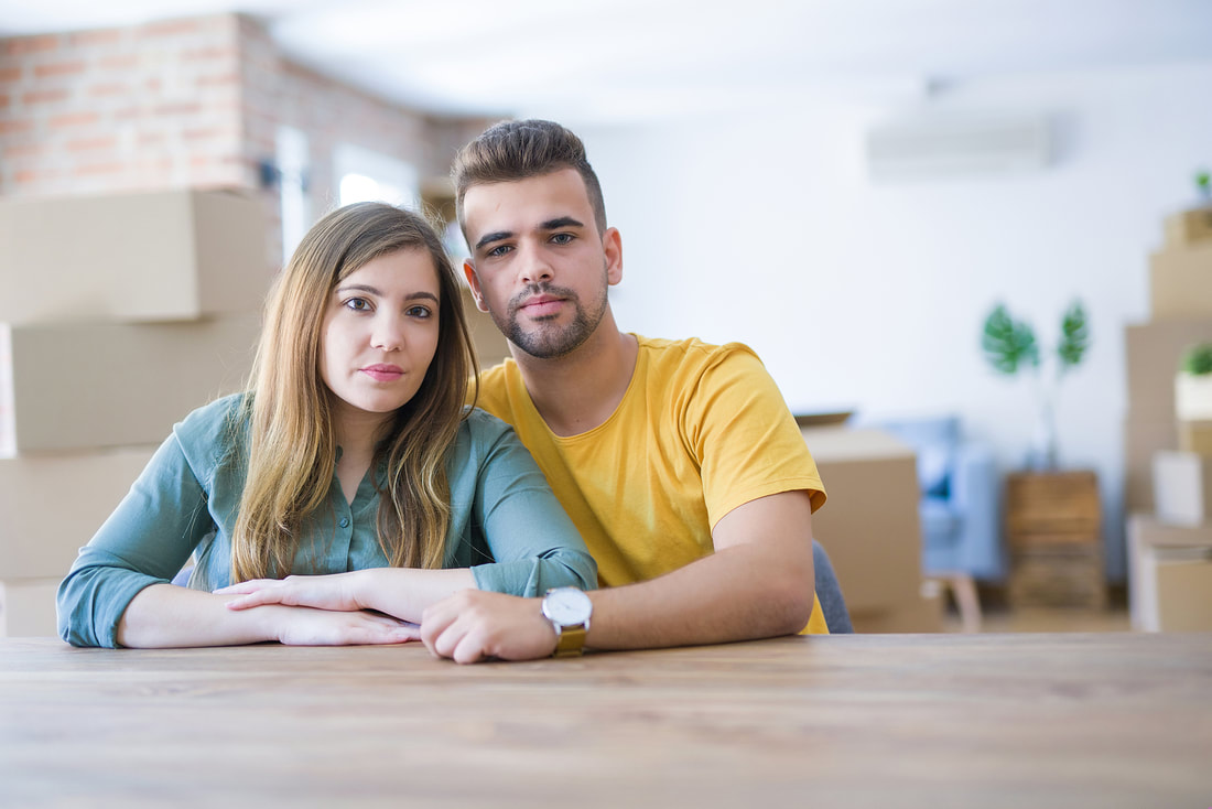Picture of couple sitting at a table with boxes all around, looking at the camera. Talk to your partner about lasting longer in bed. Then, talk with a sex therapist in Minnesota, near Minneapolis, MN and online sex therapist in Minnesota.