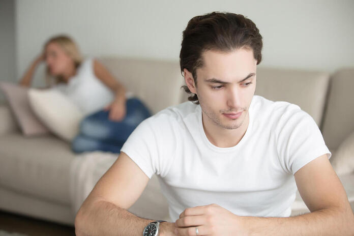 Picture of a man looking distraught while his partner is laying on the couch in the background upset about his substance abuse and sexuality consequences. Learn more from an online sex therapist in Minnesota here. We can talk about erectile dysfunction treatment Minneapolis, MN as well. Learn more here!