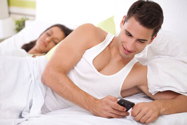 Man texting in bed smiling while his partner is asleep next to him. Creating an agreement around cheating and infidelity can help your relationship succeed long-term. Read more from a Plymouth, MN sex therapist here.  55447 | 55441 | 55442