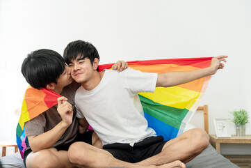 Picture of two teenage gay males, sitting cross-legged on a bed near each other, leaning in for one to kiss his partner on the cheek. They are holding a rainbow PRIDE flag, wrapping it around the two of them as they connect. Talking with your teen about sex and sexuality doesn't have to be difficult! Let our teen counselor at Sexual Wellness Institute in the Minneapolis, MN area help.