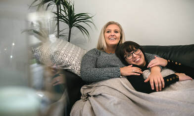 Lesbian couple watching Bridgerton, cuddling on the couch. They might be feeling physical arousal watching the sex scenes that encourage empowered female sexuality. You can get marriage counseling or sex therapy in Plymouth, MN to learn more about how masturbation for women can help you feel more connected with your partner. Get help with sex therapy in Minneapolis and couples counseling in online therapy in Minnesota here.