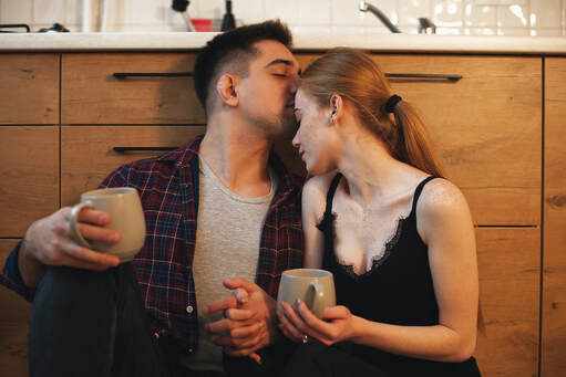Couple holding coffee cups and snuggling, sitting on the floor leaning against the cabinet in the kitchen. They are sharing a sweet moment as he kisses her forehead. They went to marriage counseling or couples therapy with a Gottman trained therapist or EFT treatment specialist to talk about low desire, low libido, mismatched desire and other sexual concerns in Plymouth, MN 55446. 